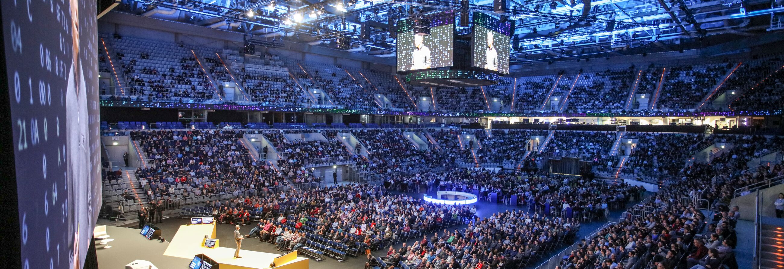 Großveranstaltung in einer voll besetzten Arena mit zentraler Bühne und Leinwänden.