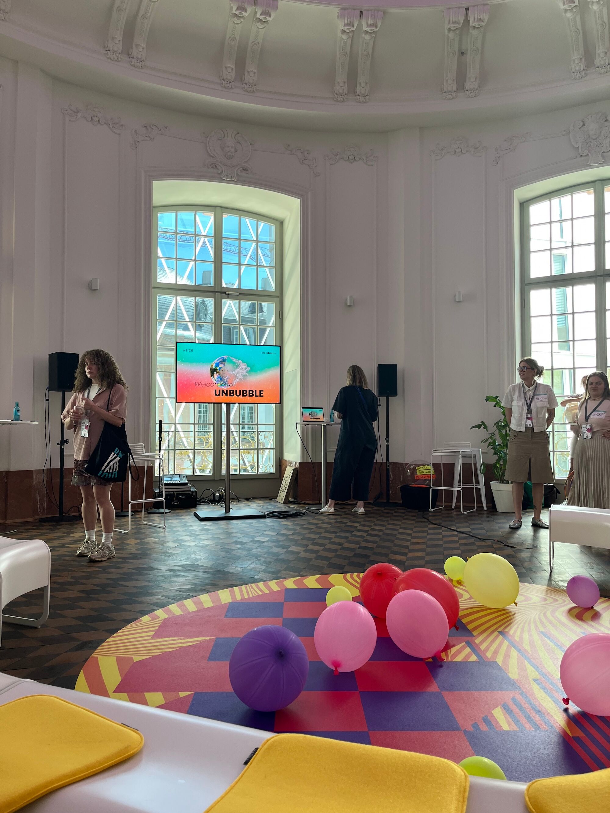 Historischer Veranstaltungsraum mit farbig gestalteter Bodenfläche, verstreuten Luftballons und einem Bildschirm mit der Begrüßung zum Format „UNBUBBLE“ vor Beginn einer interaktiven Session.