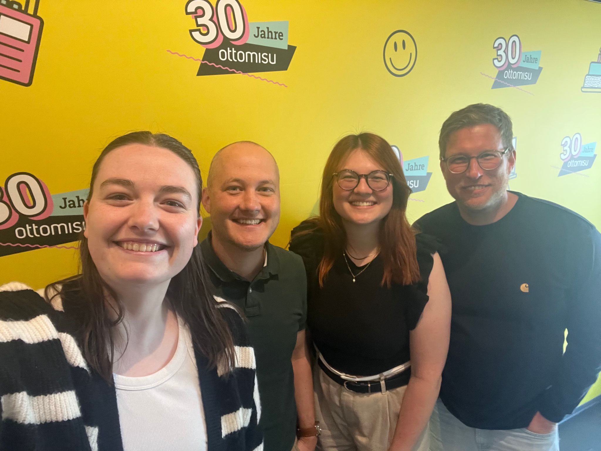 Vier Menschen stehen vor einer knallgelben Wand mit dem Aufdruck „30 Jahre ottomisu“ und posieren für ein Selfie. Alle lächeln in die Kamera. Die Wand ist mit bunten Grafikelementen und Smileys gestaltet.