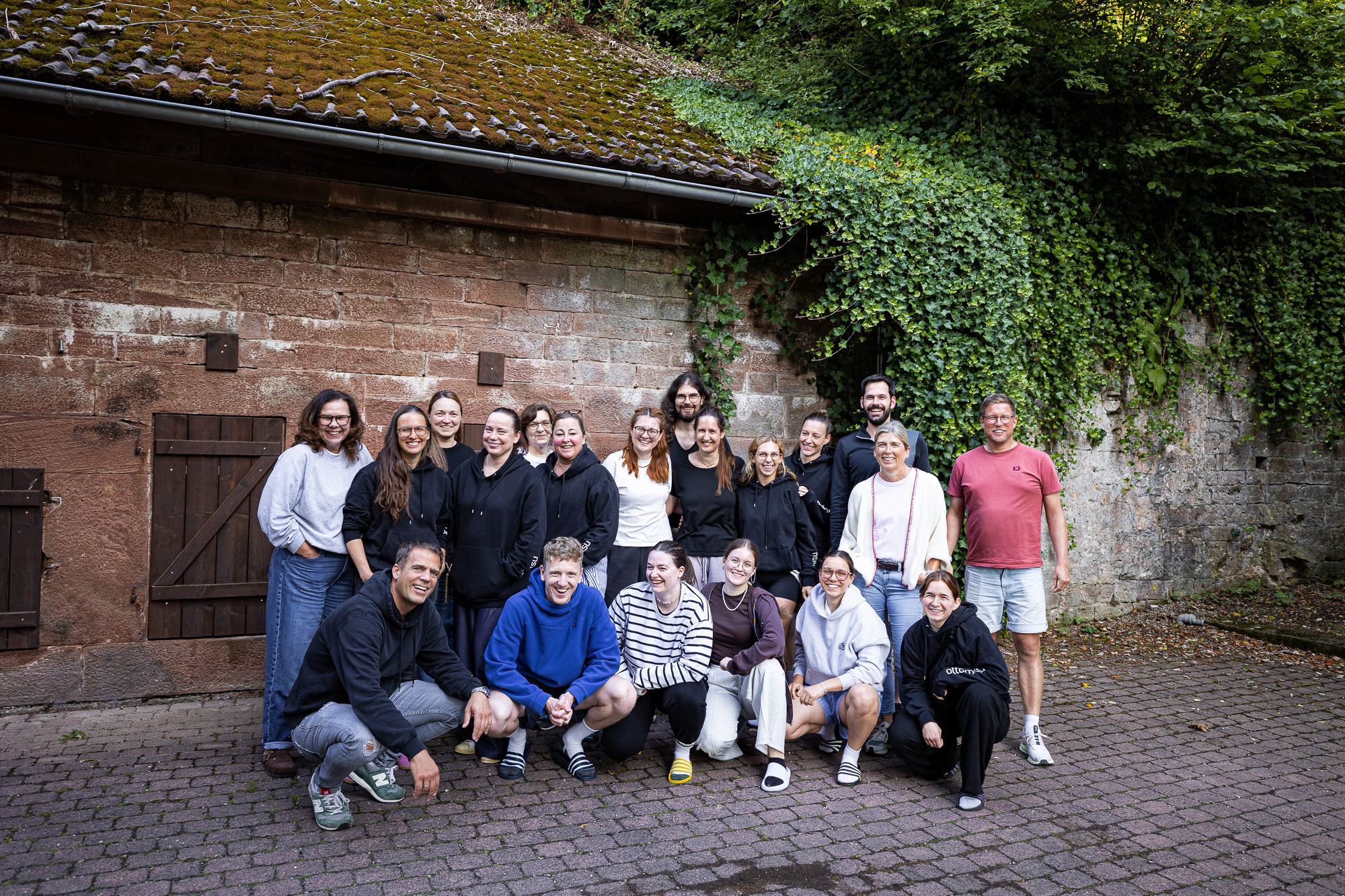 Teamfoto ottomisu vor einer mit Efeu bewachsenen Natursteinwand und einem rustikalen Gebäude mit moosbedecktem Dach. Die Personen stehen und hocken eng beieinander und strahlen eine entspannte und freundschaftliche Atmosphäre aus.