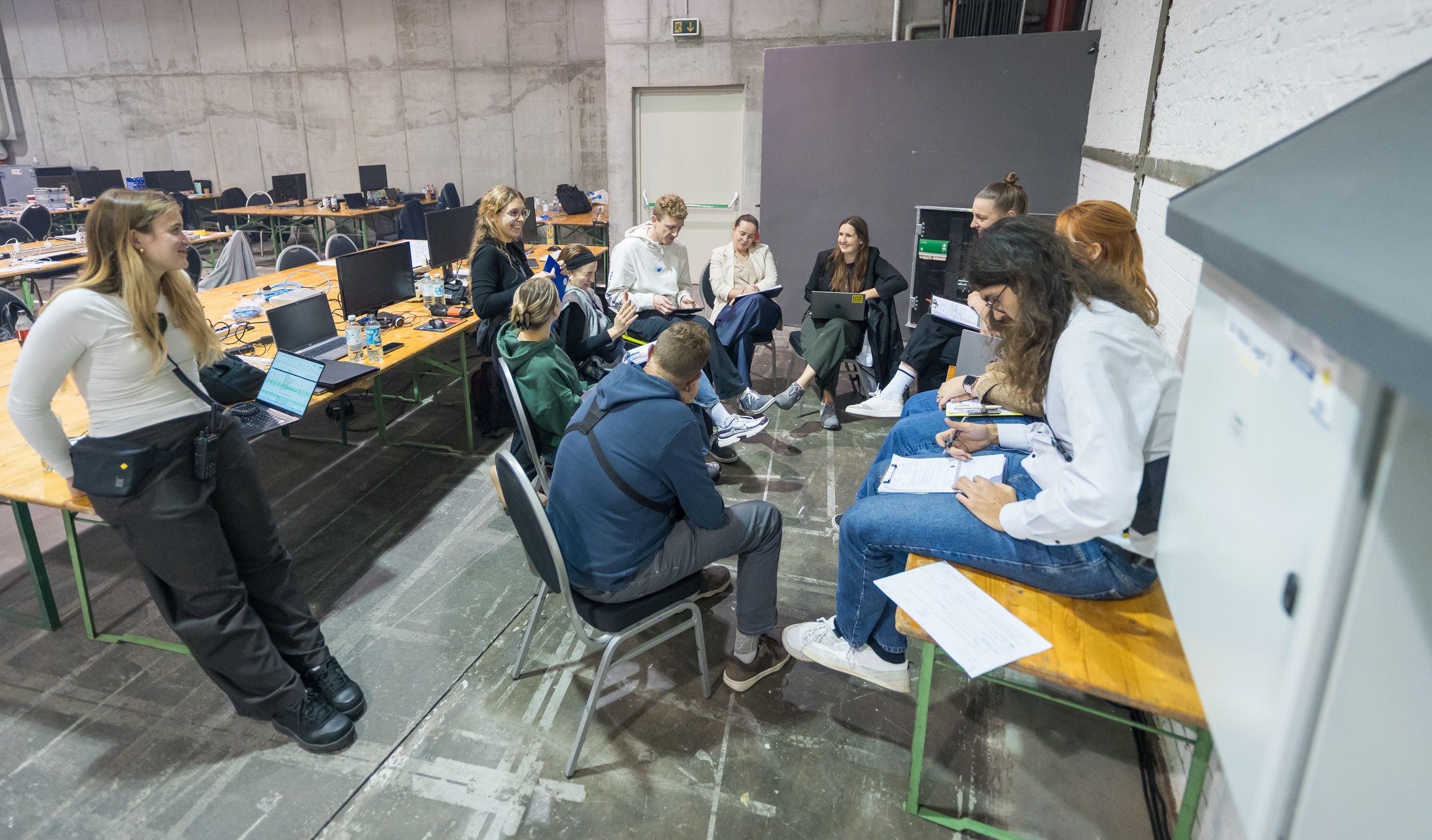 Ein Teammeeting im Backstage-Bereich einer Veranstaltung: Rund zehn Personen sitzen in einem Kreis auf Stühlen und Bänken, sprechen und machen sich Notizen. Im Hintergrund stehen Tische mit Laptops, Wasserflaschen und Technikmaterial.