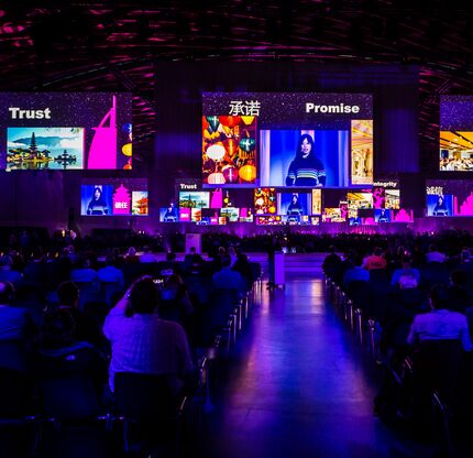 Blick in einen großen Eventsaal mit Bühne, mehreren Leinwänden und farbenfroher Lichtinszenierung.