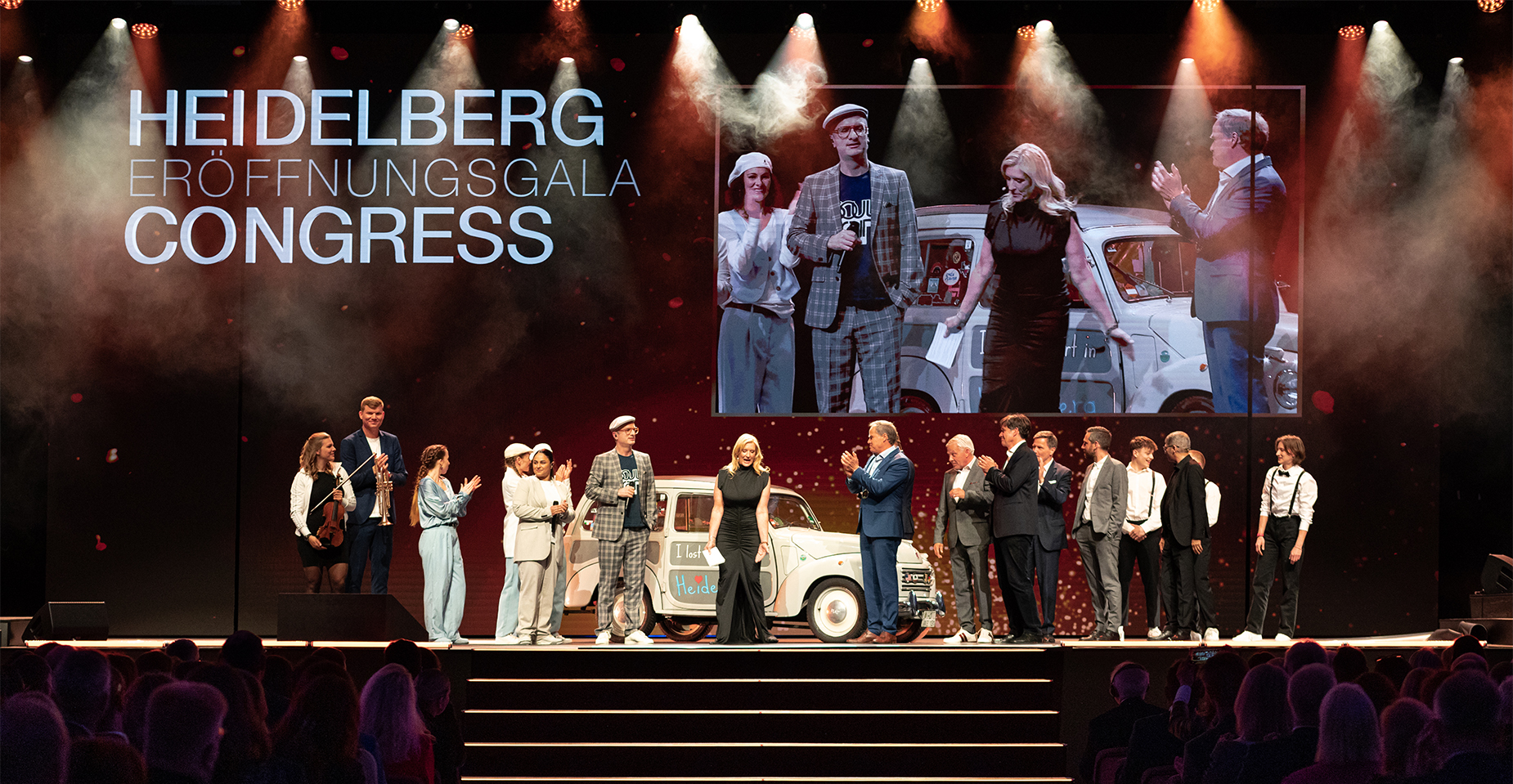 uf einer Bühne stehen etwa 15 Personen nebeneinander. Im Hintergrund ist ein großes projiziertes Gruppenfoto zu sehen, darüber steht ‚Heidelberg Congress‘. Die Szene ist feierlich beleuchtet.