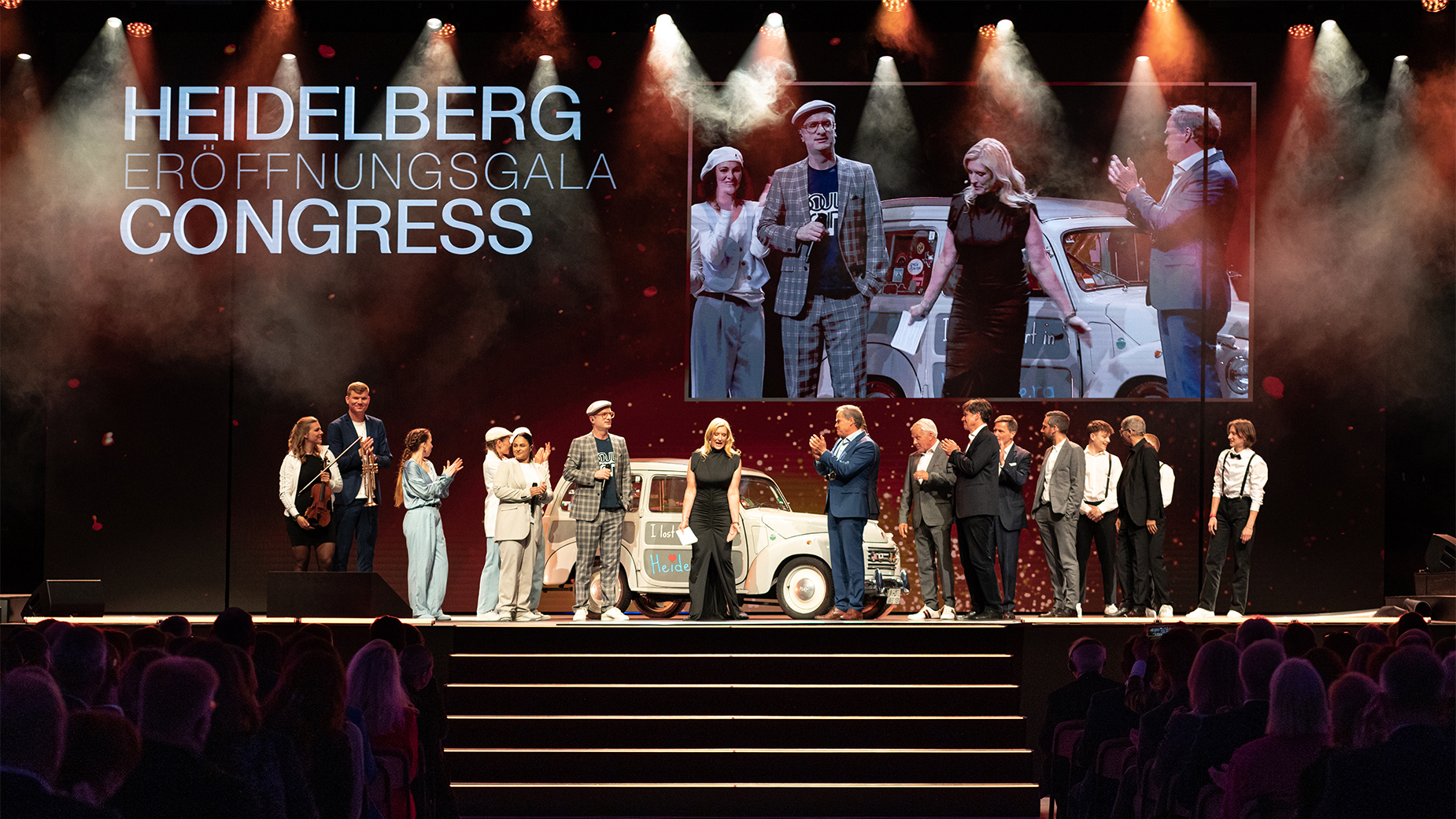 uf einer Bühne stehen etwa 15 Personen nebeneinander. Im Hintergrund ist ein großes projiziertes Gruppenfoto zu sehen, darüber steht ‚Heidelberg Congress‘. Die Szene ist feierlich beleuchtet.