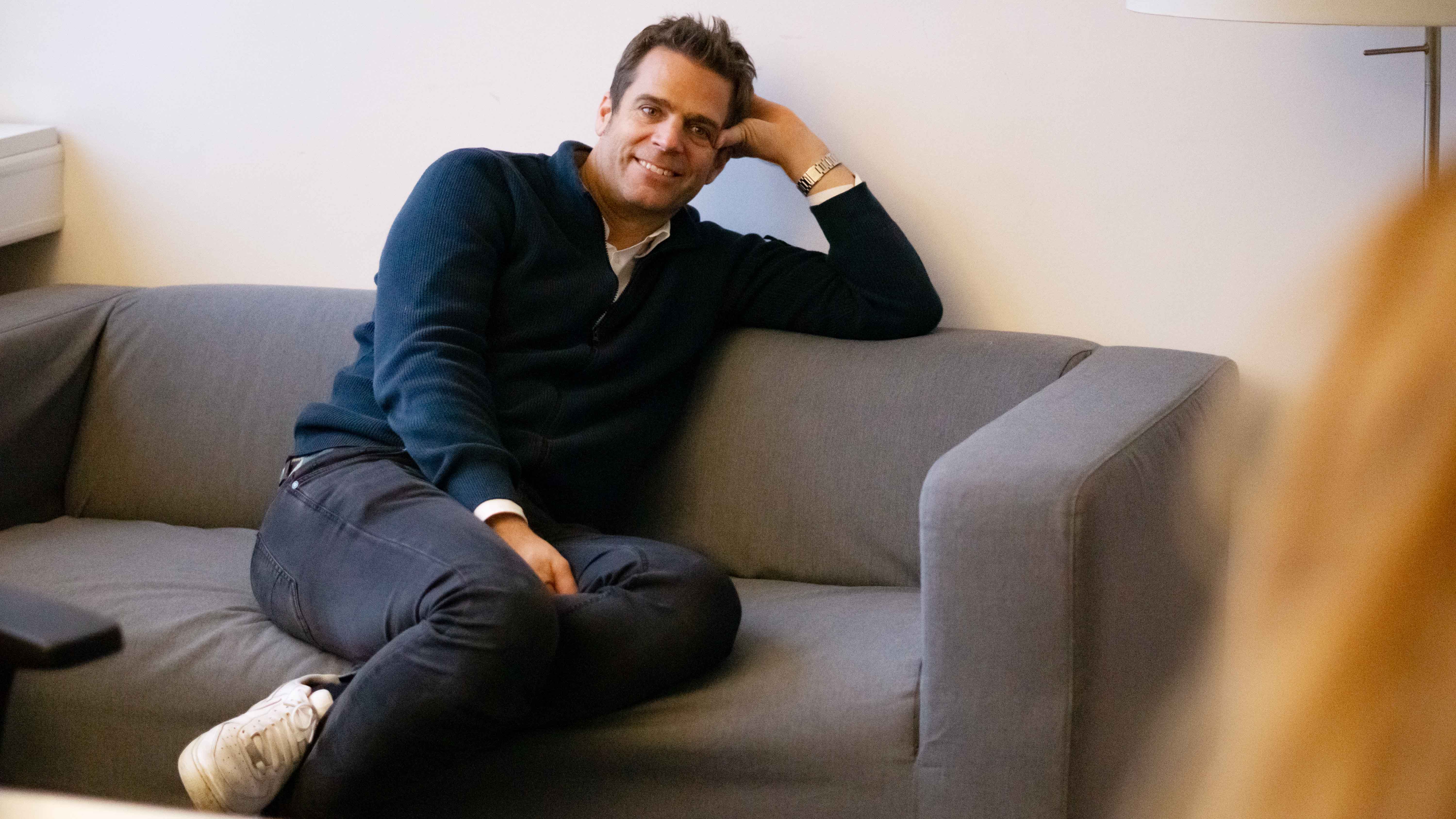 ottomisu CEO Jörn Huber sitzt auf einem grauen Sofa im Büro und führt ein Interview mit Kolleg*innen.