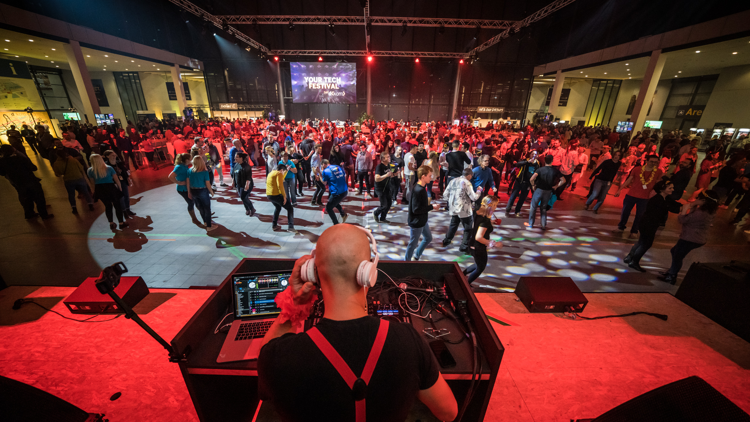 Im Vordergrund befindet sich ein DJ mit DJ-Pult, der auf die Tanzfläche mit den sich amüsierenden Besucher*innen blickt.