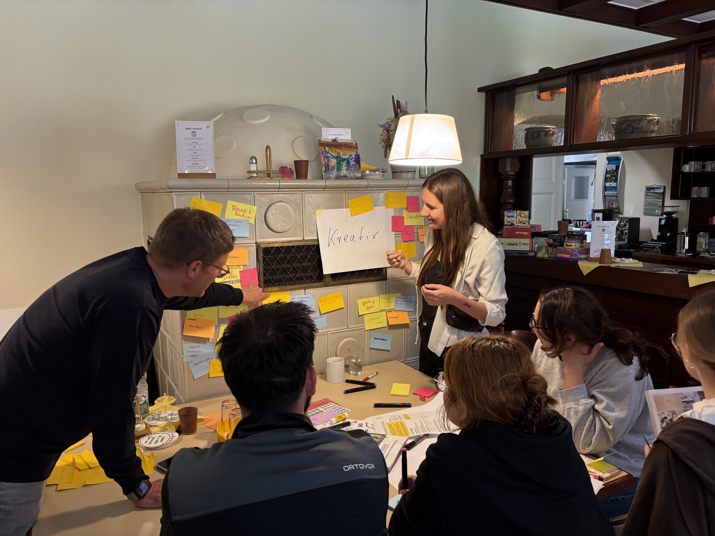 Eine Frau moderiert eine Kreativsession an einem Tisch, der mit bunten Post-its bedeckt ist. Im Hintergrund ein alter Kachelofen, an dem ein großes Papier mit der Aufschrift „Kreativ“ hängt. Die Gruppe hört aufmerksam zu.