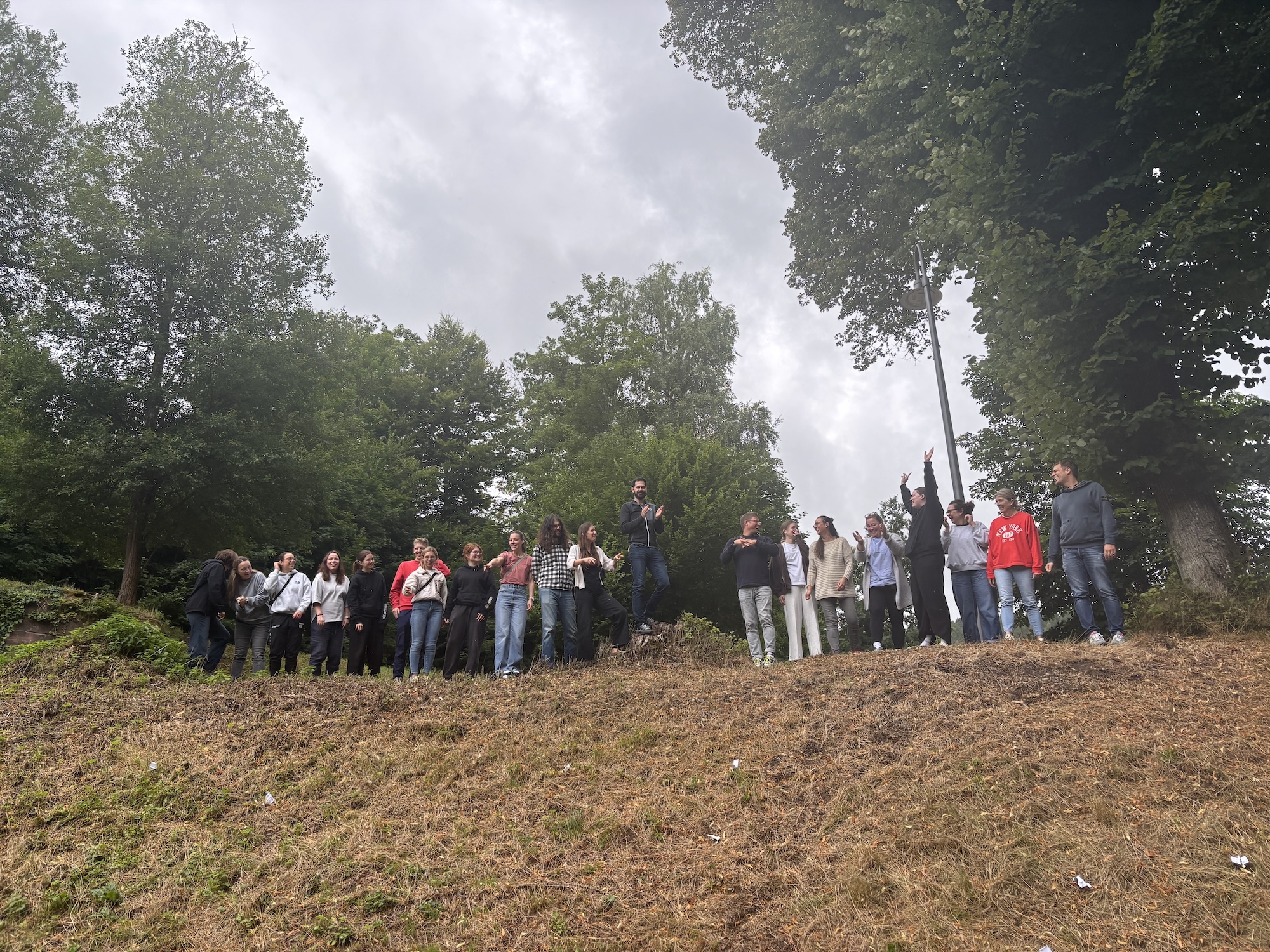Eine Gruppe von Menschen steht auf einem Hügel vor bewaldetem Hintergrund. Einige lachen und unterhalten sich, andere blicken neugierig nach vorne. Der Himmel ist wolkenverhangen.