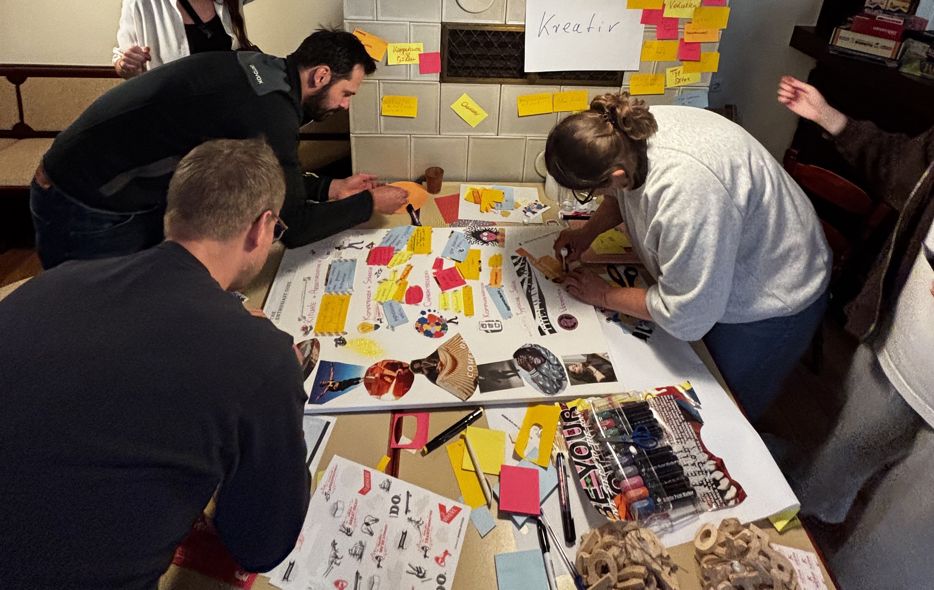 Mehrere Personen arbeiten gemeinsam an einem großen Tisch an einer Collage. Es liegen Scheren, Stifte, Kleber, ausgeschnittene Bilder und Post-its bereit. An der Wand hängt ein Schild mit der Aufschrift „Kreativ“.