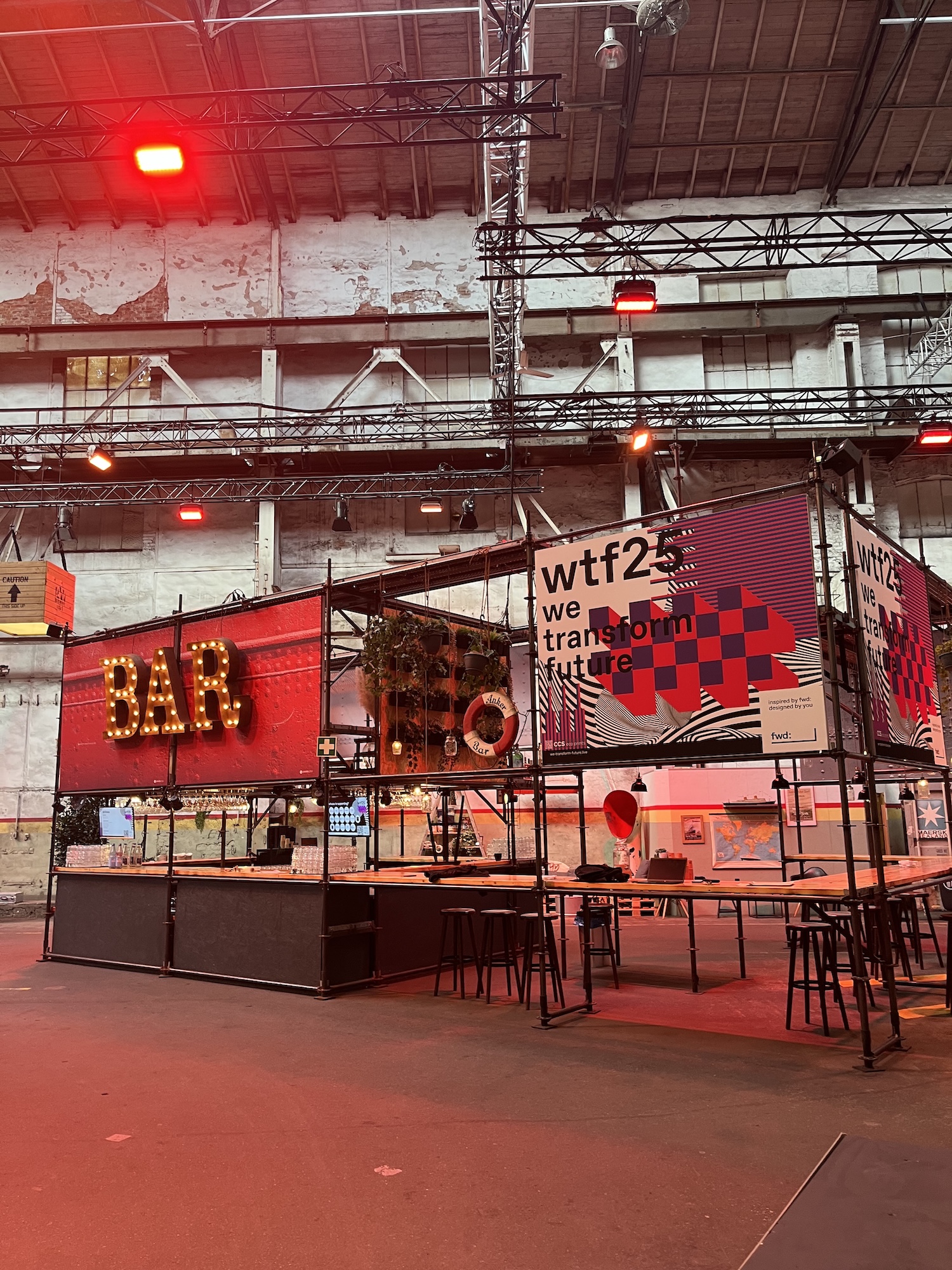 Stylisch inszenierte Bar im Innenbereich der Veranstaltungshalle mit rot leuchtendem „BAR“-Schriftzug und dem Branding von wtf25. Der industrielle Look wird durch Gerüste, Lichtinstallationen und Dekoelemente unterstrichen.