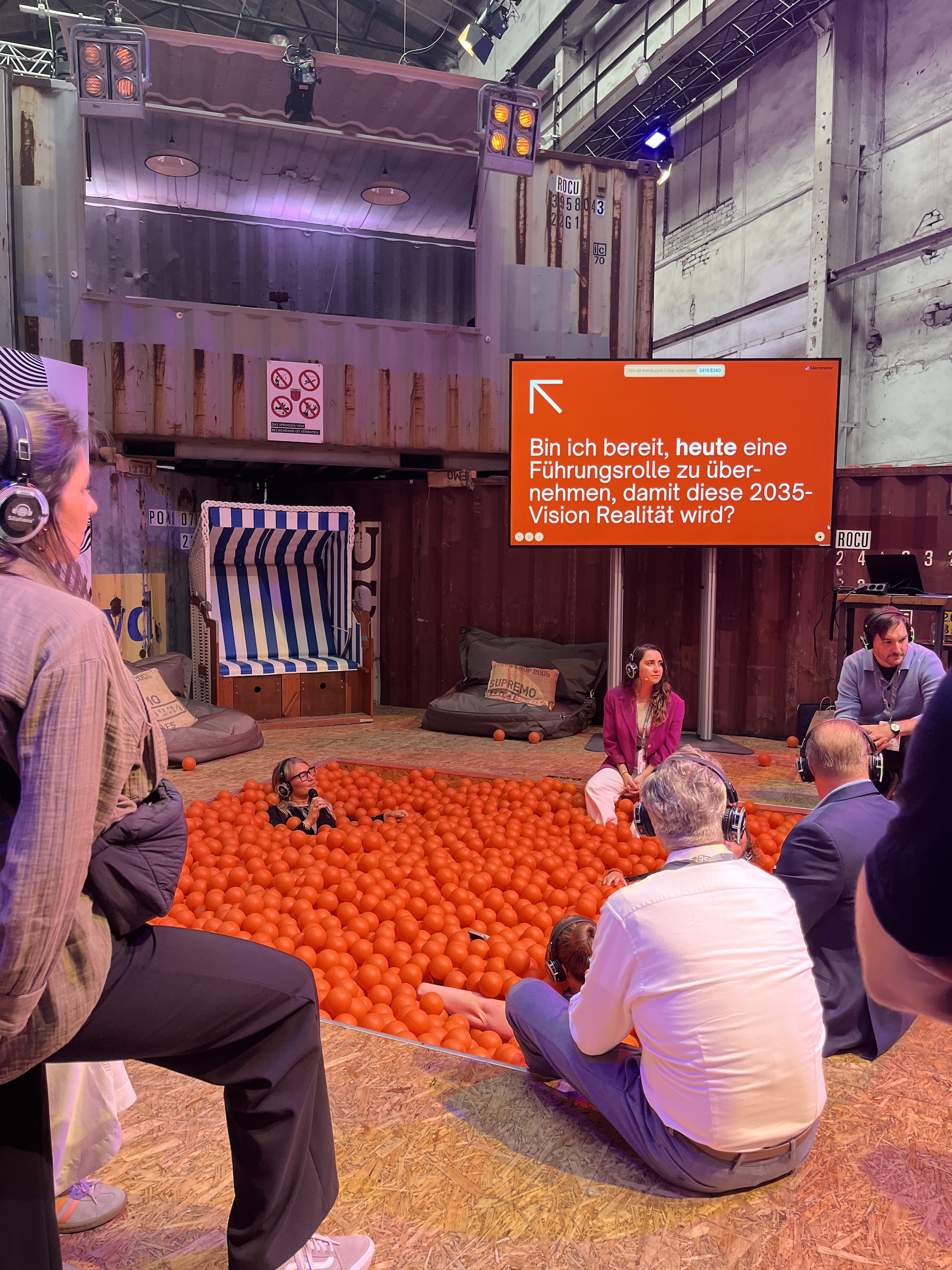 Kleine Gesprächsrunde mit Kopfhörern in der „interaction area“ von wtf25, teilweise im orangen Bällebad sitzend. Auf dem Screen die Frage: „Bin ich bereit, heute eine Führungsrolle zu übernehmen, damit diese 2035-Vision Realität wird?“