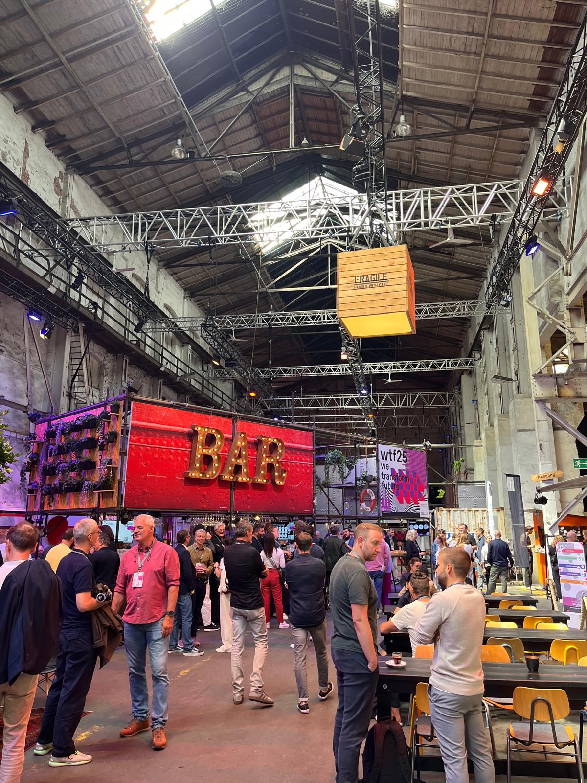 Lebhafter Barbereich auf dem wtf25-Event mit vielen Gästen im Gespräch. Im Vordergrund ist ein leuchtender „BAR“-Schriftzug zu sehen, darüber das Eventbranding und Industriekulisse mit hoher Decke.