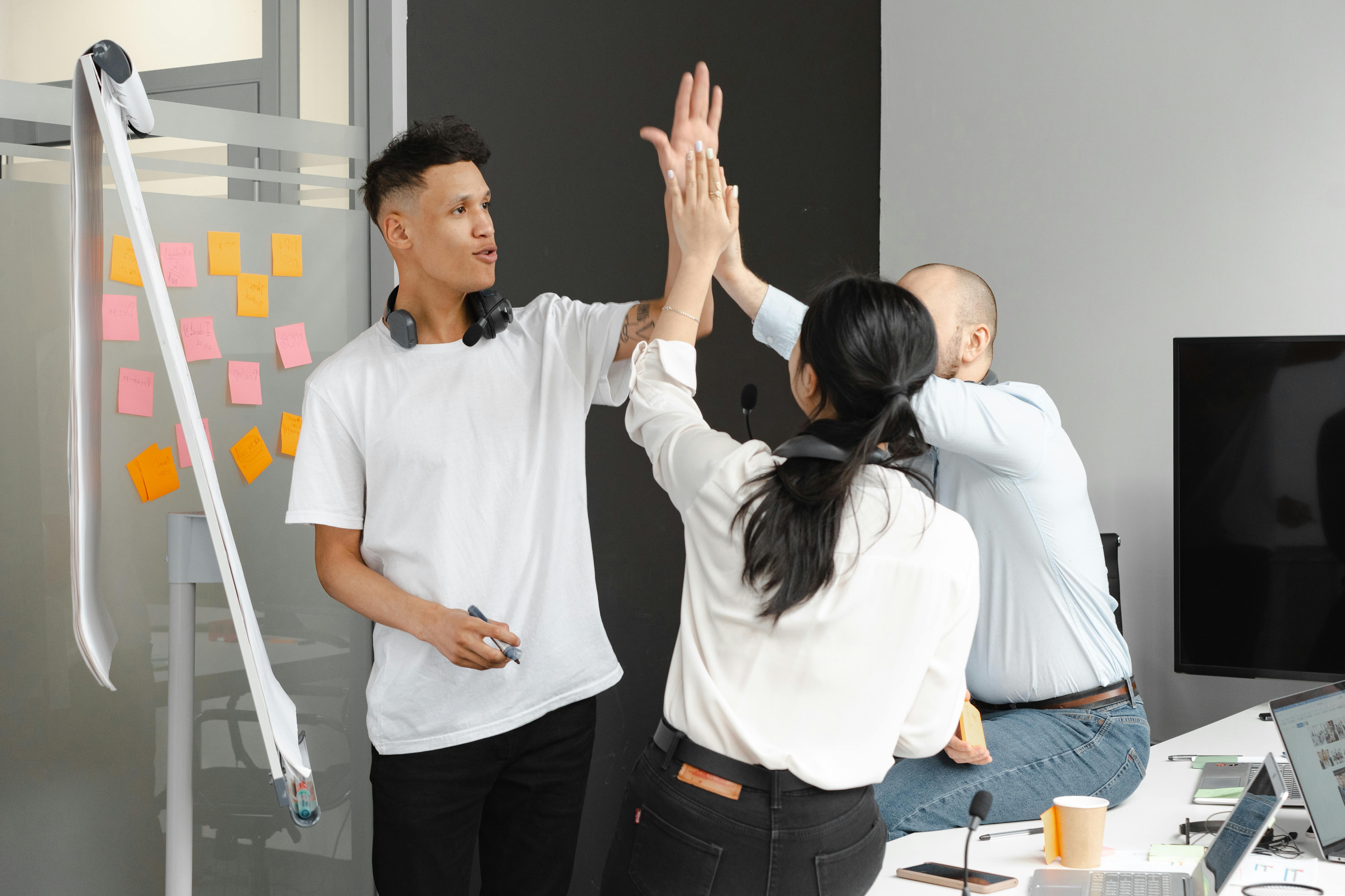 Drei Kolleg:innen geben sich im Büro ein High-Five und feiern einen Erfolg. Im Hintergrund ein Flipchart mit bunten Post-its, ein Laptop und ein Bildschirm. Die Stimmung ist motivierend und dynamisch.