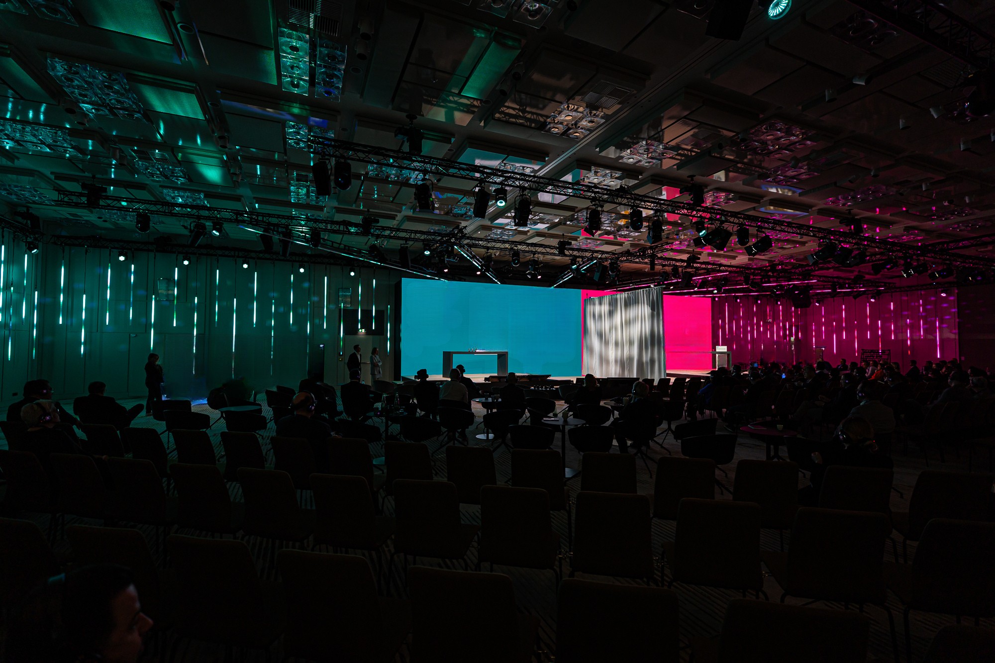 Eventlocation mit Konferenzbestuhlung und zweigeteiltem Bühnenbereich, Menschen im Gespräch vor Veranstaltungsbeginn.