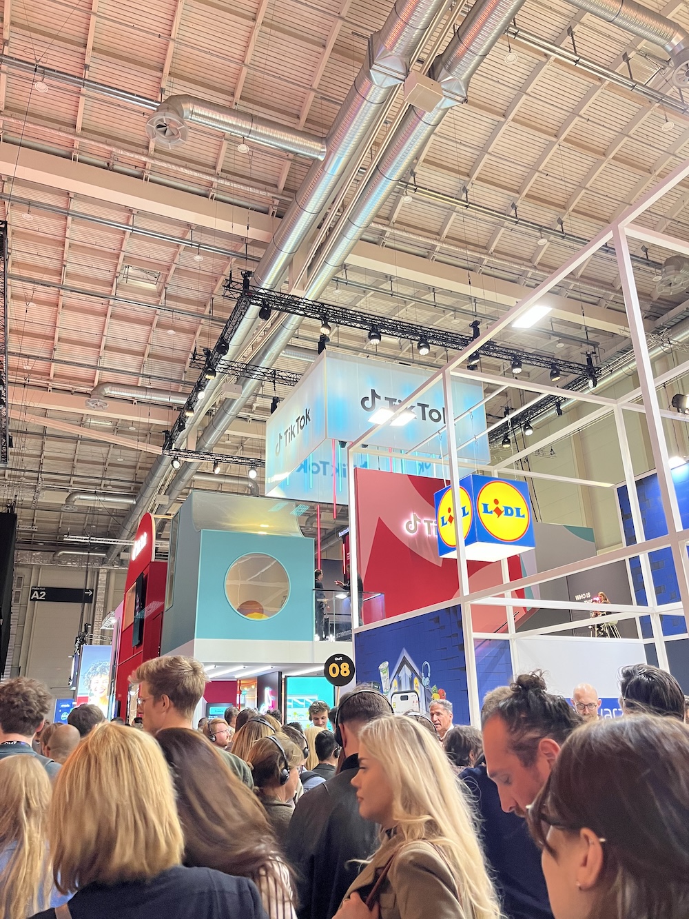 Messehalle mit hohen, auffälligen Markenbauten von TikTok und Lidl, große Menschenmenge davor, buntes Lichtambiente.