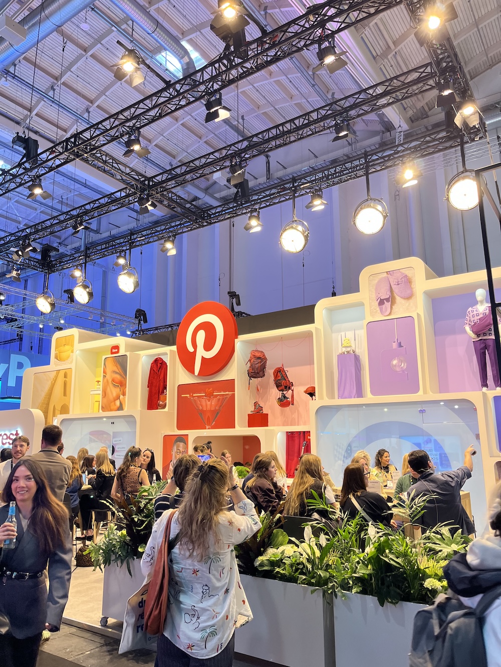 Pinterest-Messestand mit Regalwänden, Produkten und Pflanzen-Elementen, viele Besucher*innen in Gesprächen davor.