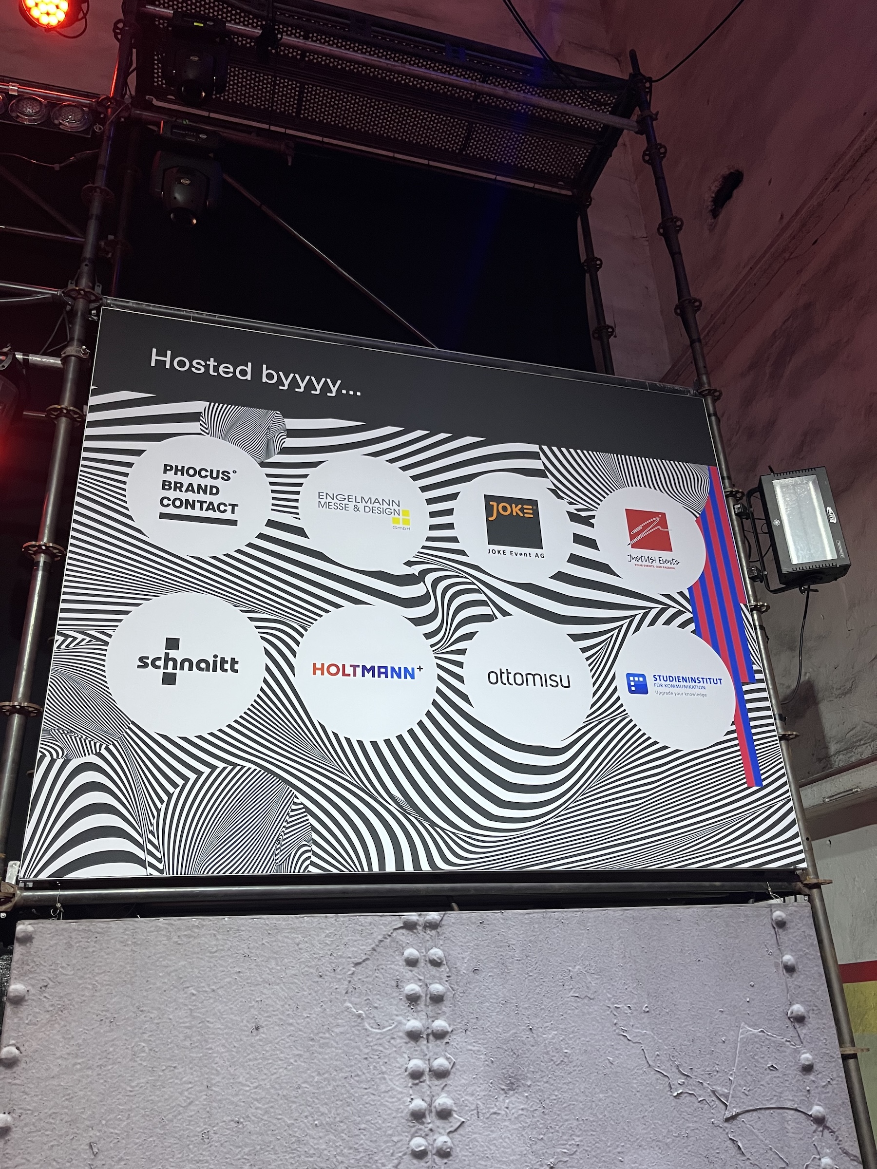 Sponsorenwand des Events mit dem Schriftzug „Hosted byyyy...“ und Logos von Partnerunternehmen wie Phocus Brand Contact, Engelmann Messe & Design, JOKE Event AG, ottomisu, u.a., auf psychedelischem Schwarz-Weiß-Hintergrund.