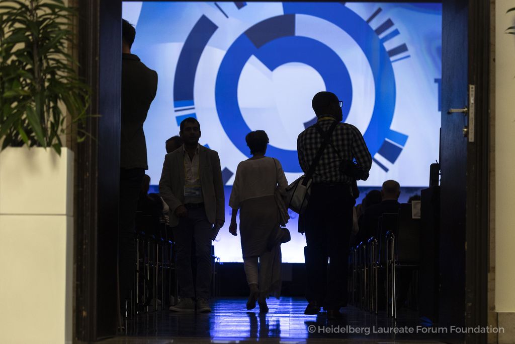 Teilnehmende des Heidelberg Laureate Forums betreten einen verdunkelten Vortragssaal mit großer LED-Wand im Hintergrund. Die Lichtstimmung ist zurückhaltend, die Silhouetten der Personen sind im Gegenlicht sichtbar.