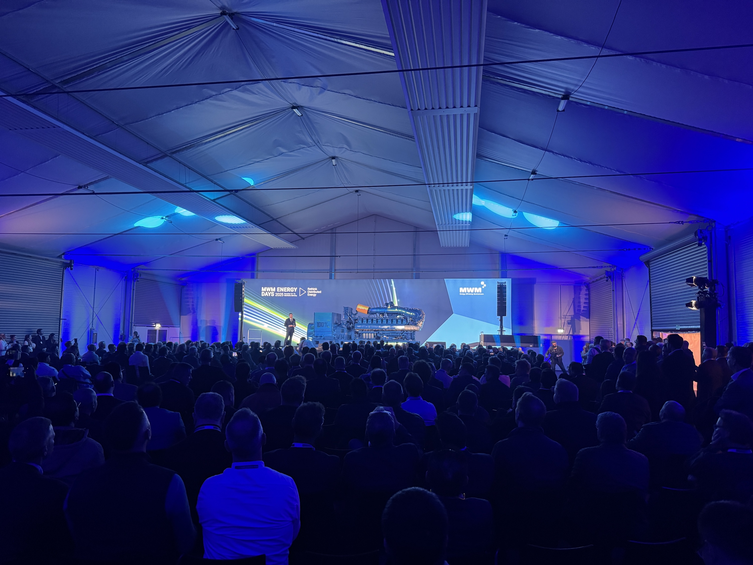 Blick ins große Eventzelt der MWM Energy Days mit Publikum und Bühne. Im Zentrum steht ein Sprecher vor der beleuchteten Motoranlage und LED-Wand mit den Slogans „Rethink Distributed Energy“ und „MWM“.
