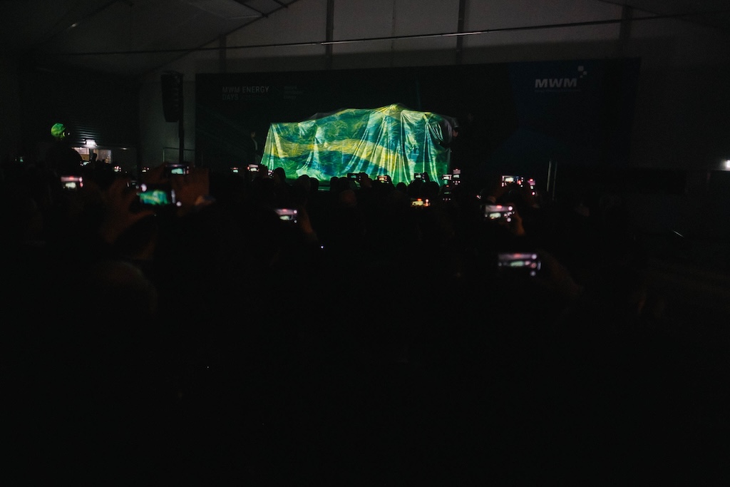 Publikum fotografiert gespannt das verhüllte Objekt auf der Bühne bei gedimmtem Licht. Zahlreiche Smartphones leuchten im abgedunkelten Raum.