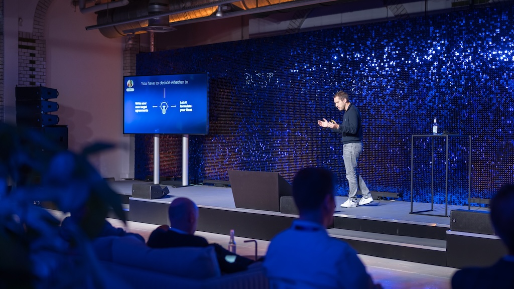 Speaker auf der Bühne vor einer schimmernden, blauen Glitzerwand. Auf dem Screen daneben eine Folie zum Thema KI: „You have to decide whether to… Write your own target agreements / Let AI formulate your ideas“.