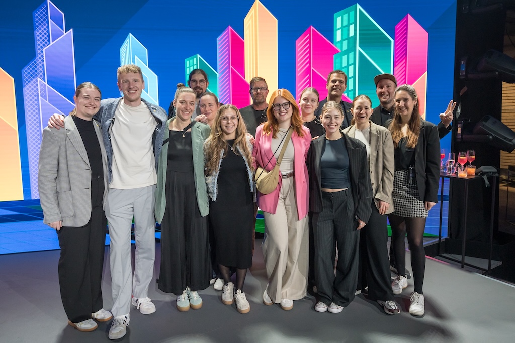 Teamfoto vor bunter LED-Kulisse mit stilisierten Hochhäusern. Ein fröhliches, diverses Team posiert nach dem Event auf der Bühne.