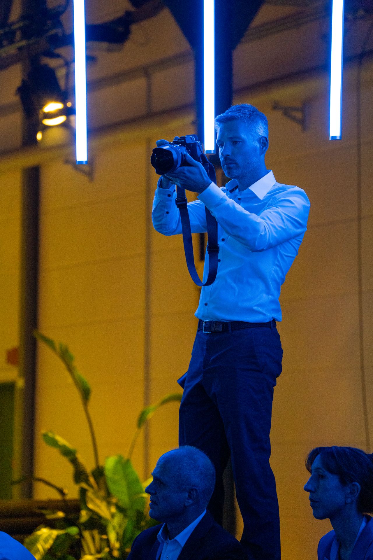 Photographer in a white shirt stands at an event under blue lighting, capturing images with a camera.