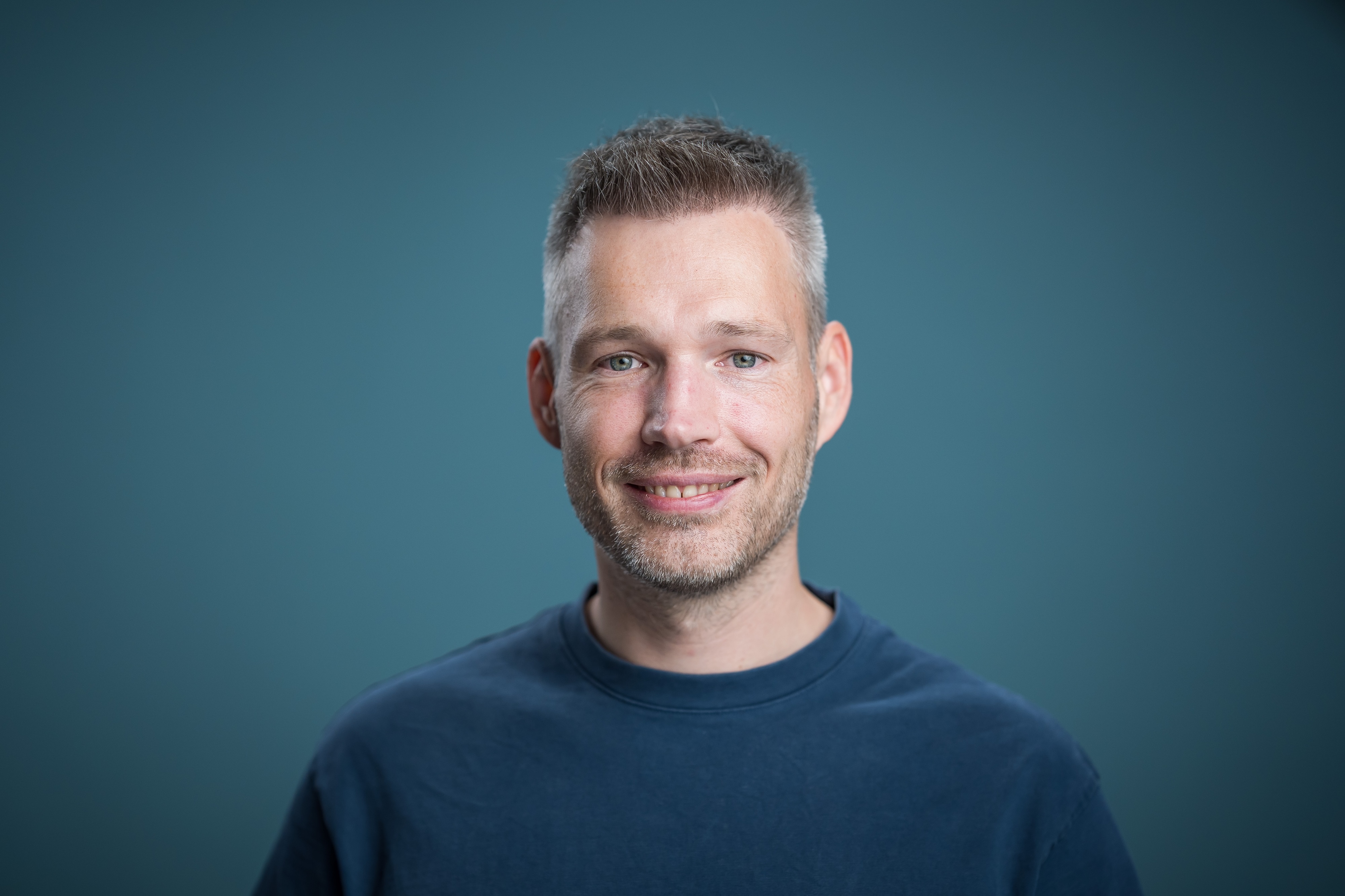 Portrait of a smiling man wearing a dark shirt against a neutral blue background.