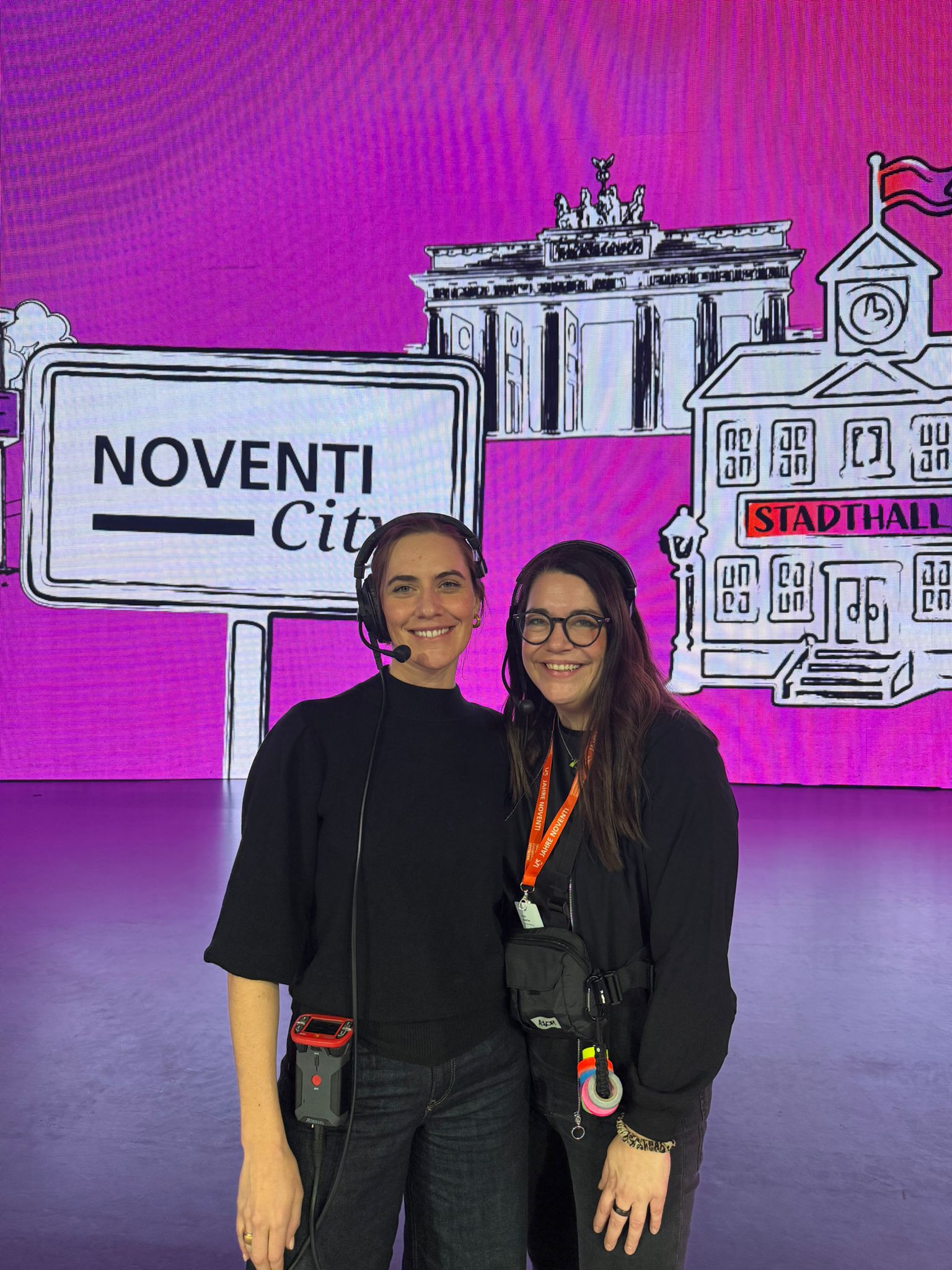 Event moderation team wearing headsets in front of a corporate event stage in “NOVENTI City” with LED visuals and stage backdrop