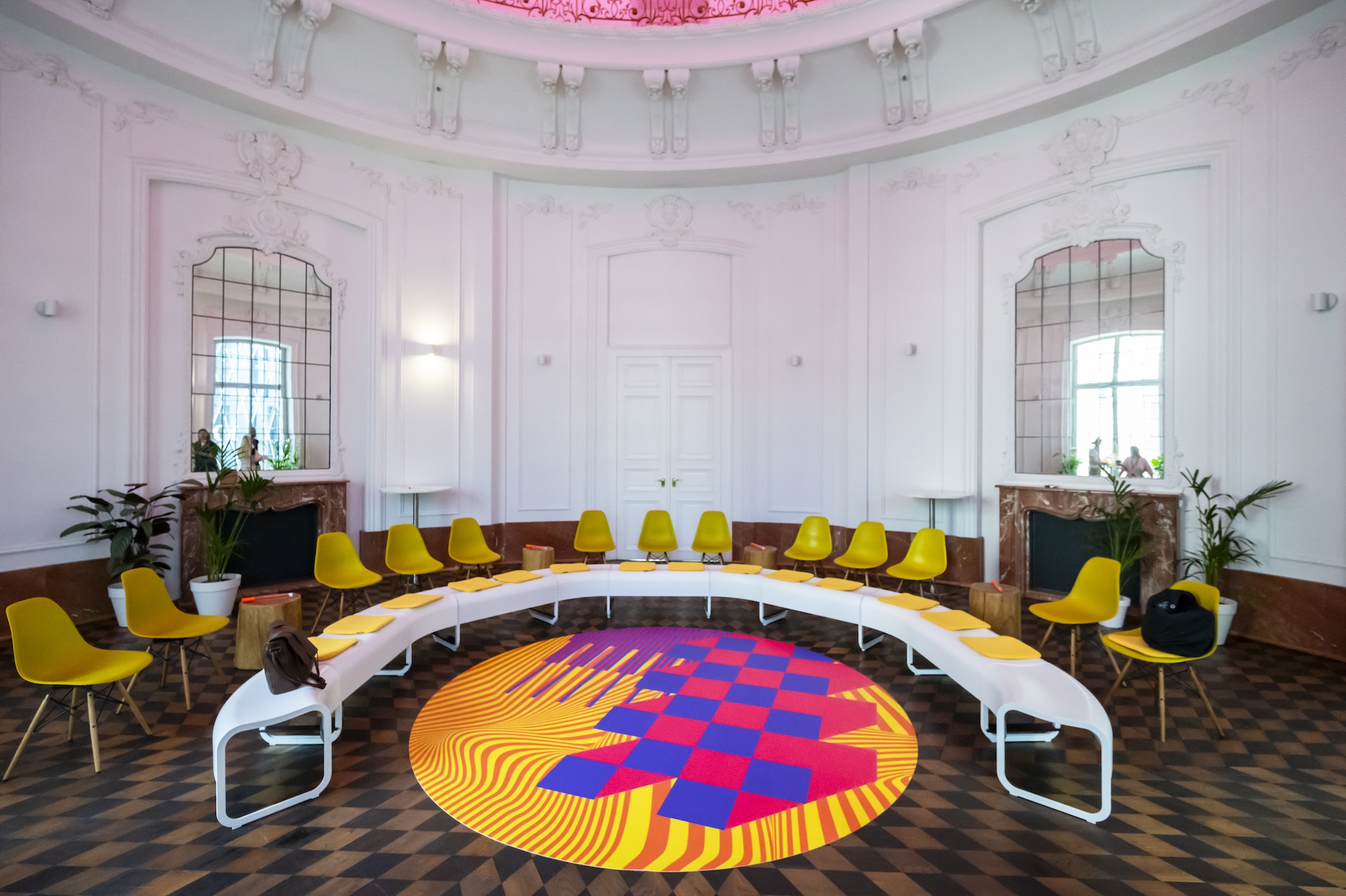 Halbkreisförmig angeordnete weiße Sitzbänke und gelbe Stühle in einem hellen historischen Saal mit hohen Fenstern, Spiegeln und einem farbigen Bodenelement in der Mitte als zentralem Treffpunkt für Workshops und Dialogformate.