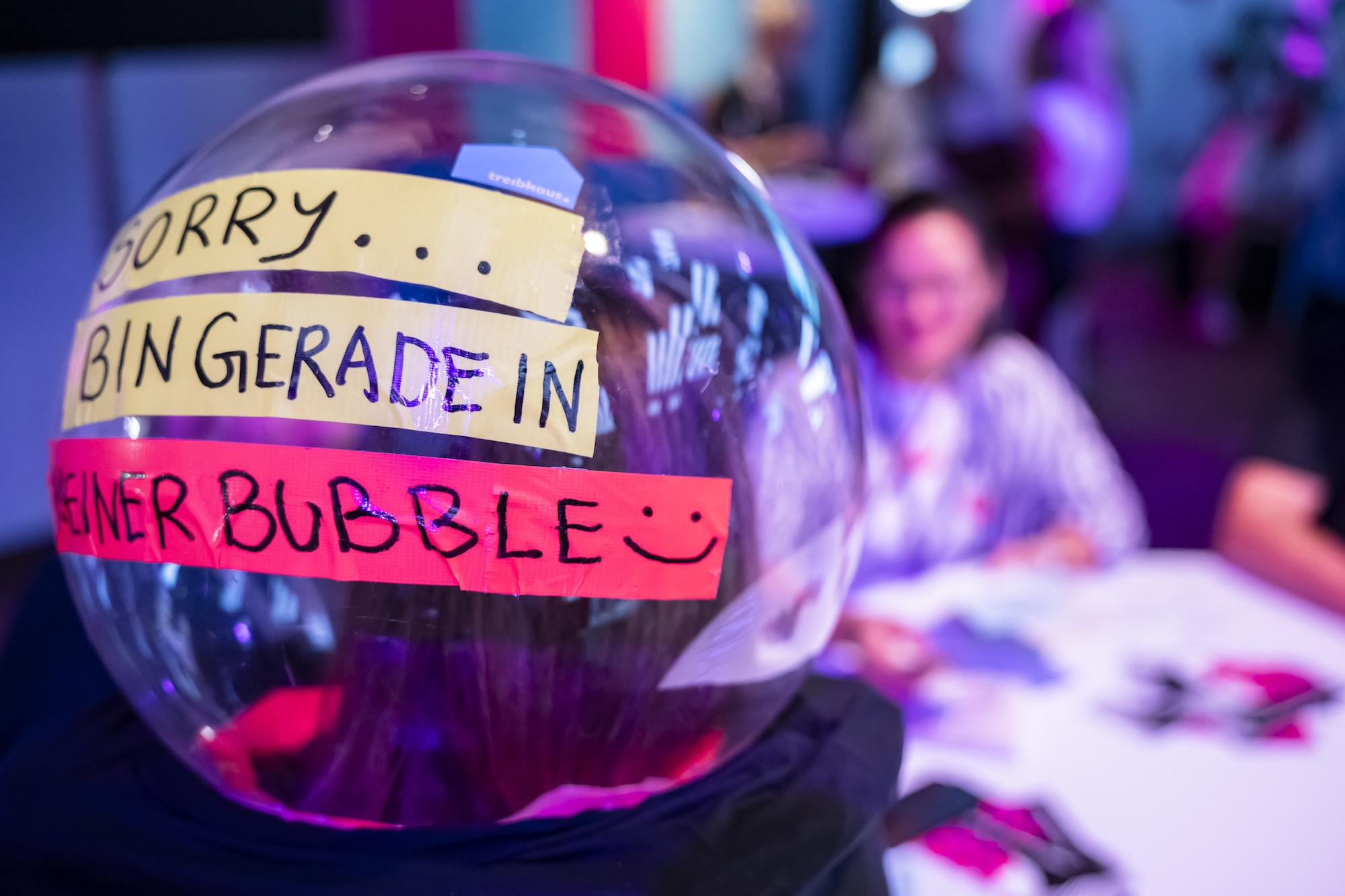 Transparente Kunststoffkugel mit der Aufschrift „Sorry, bin gerade in einer Bubble“ vor einem unscharfen Workshop-Setting mit Tischen, Teilnehmenden und farbiger Beleuchtung.