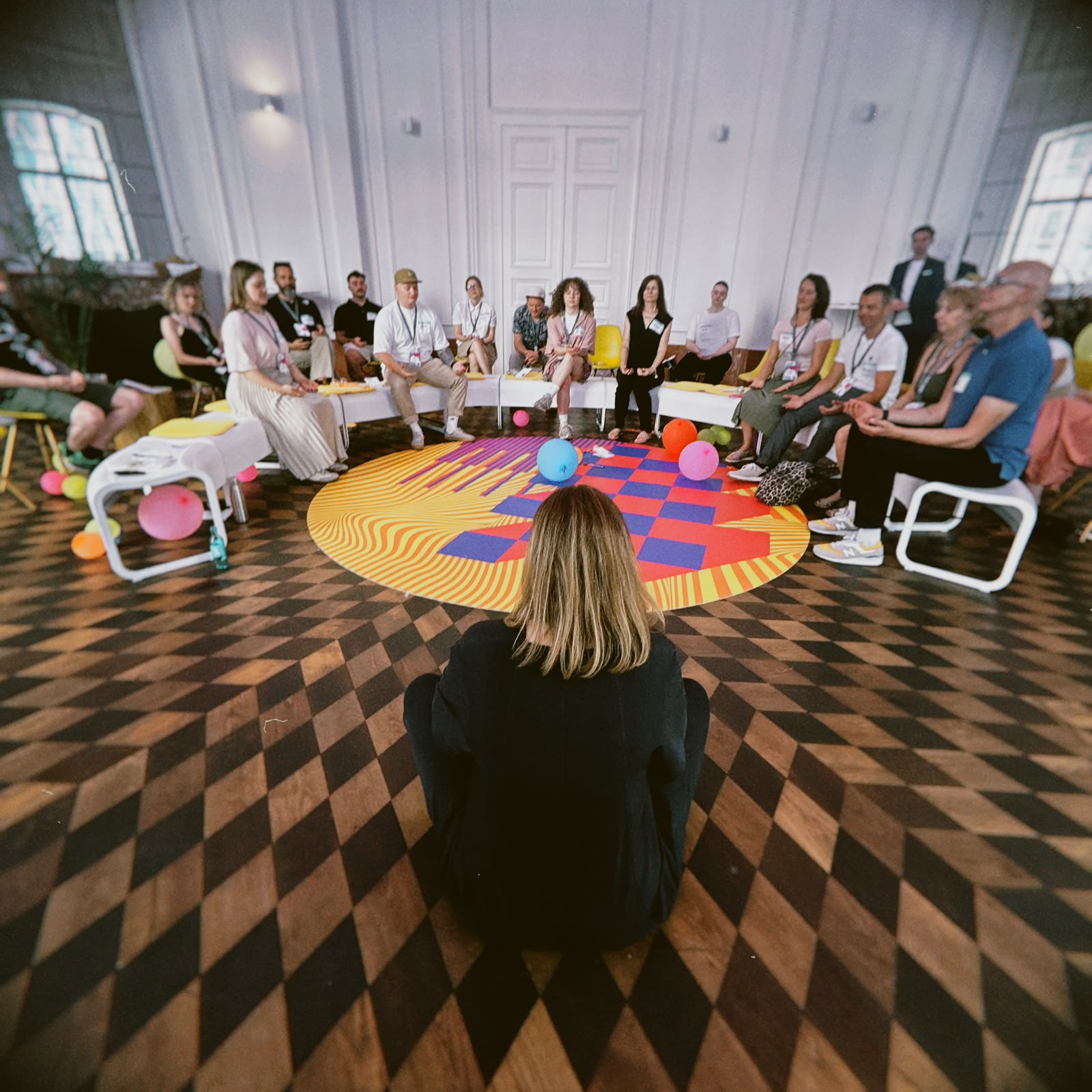Teilnehmende sitzen im Kreis rund um eine großflächige farbige Bodeninstallation und tauschen sich im Rahmen eines moderierten Workshop- und Dialogformats aus.