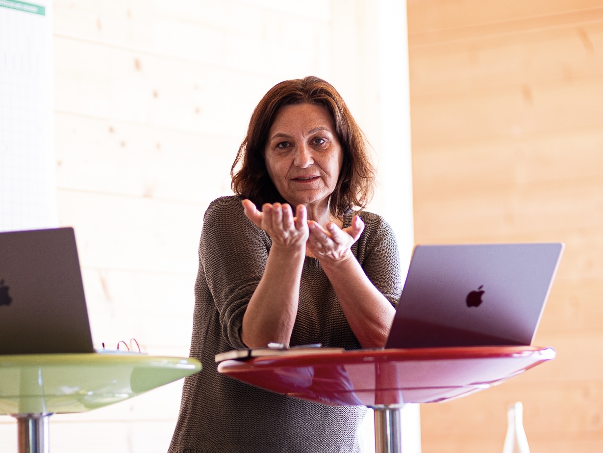 Christine explains a topic during a workshop, gesturing between two standing tables with open laptops.