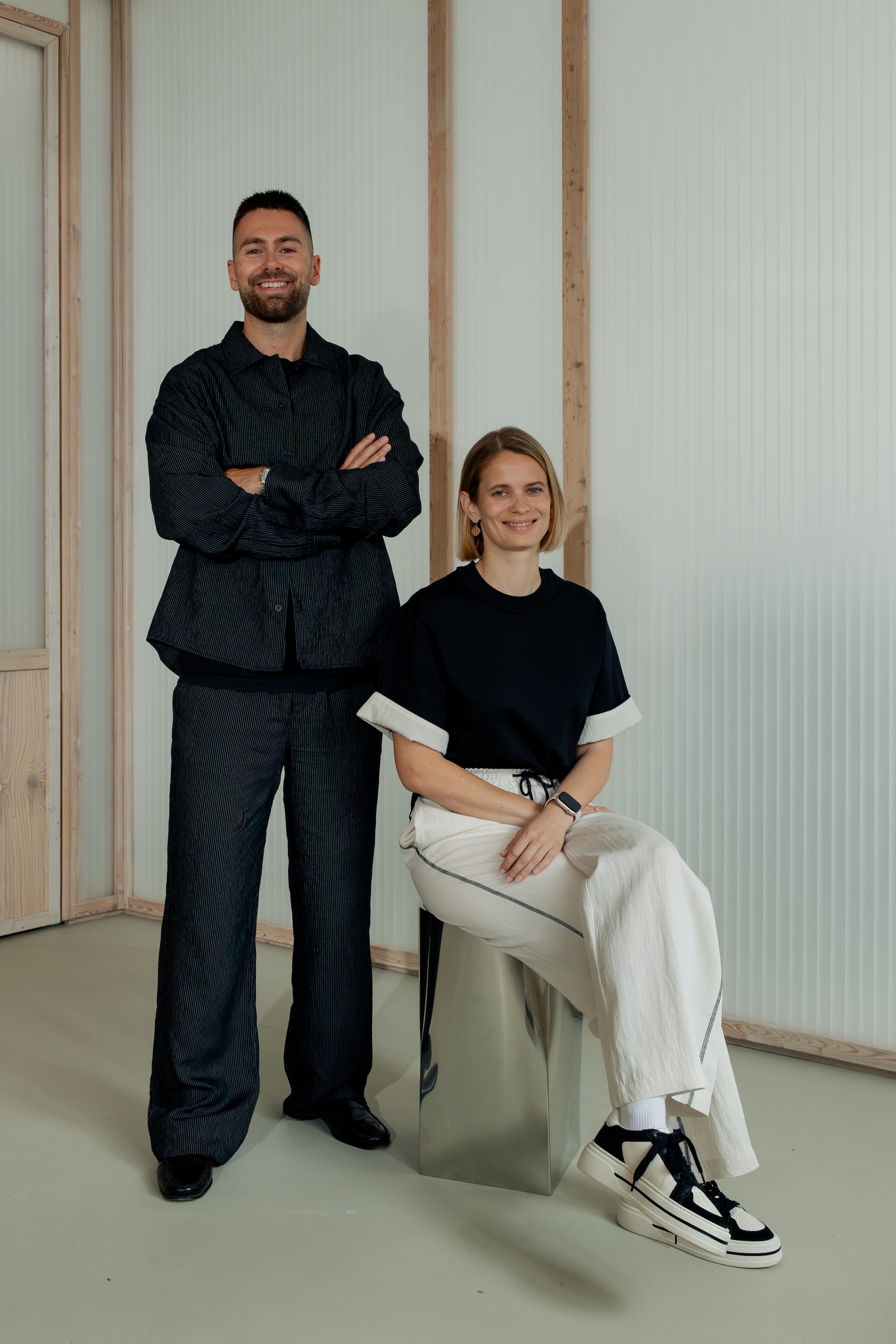Two people in dark and light casual clothing in a modern interior. One person is standing with folded arms while the other is seated on a metallic cube.