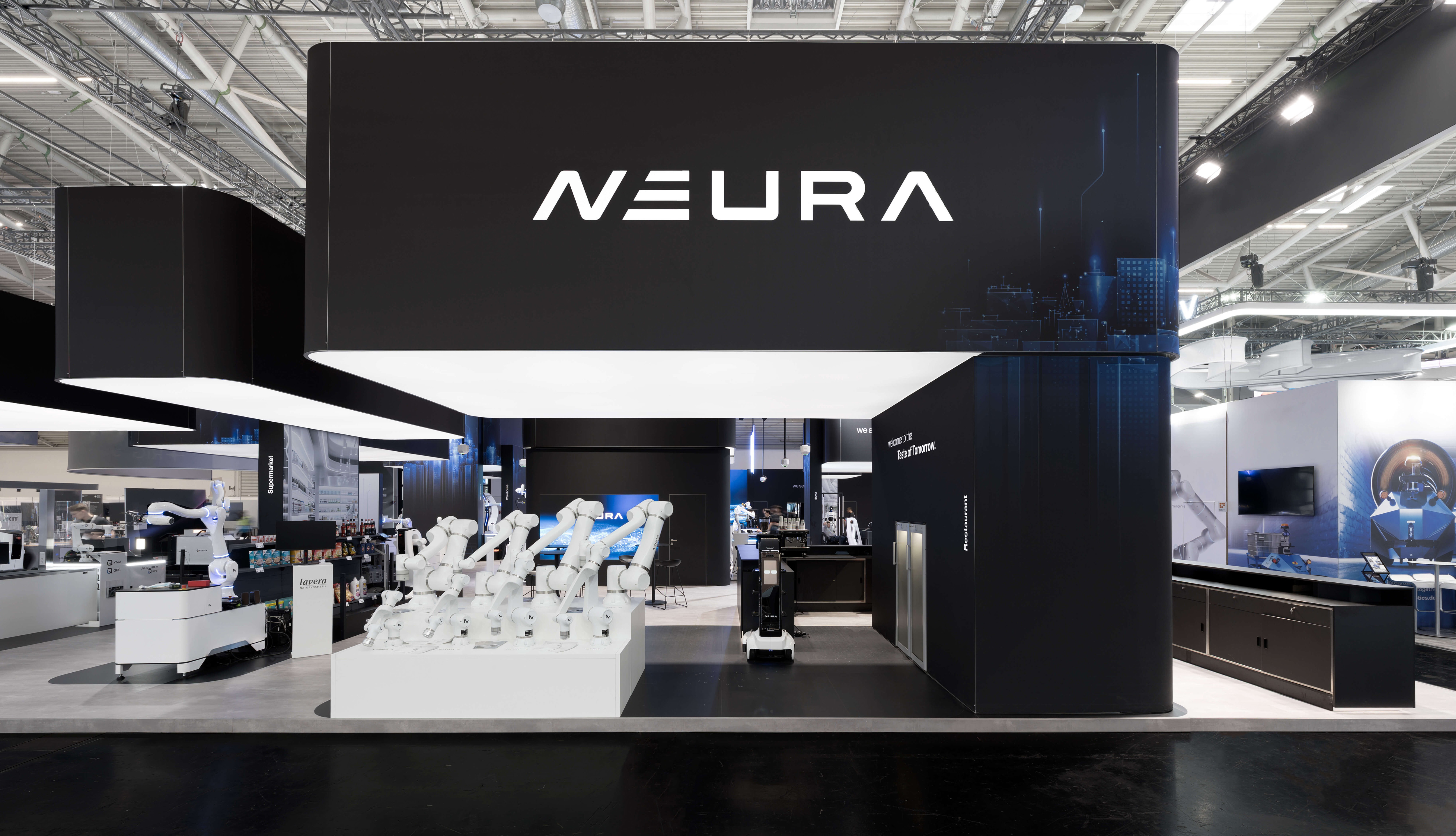 Large trade show booth featuring the “NEURA” logo, dark display structures, and robotic systems exhibited inside a convention hall.